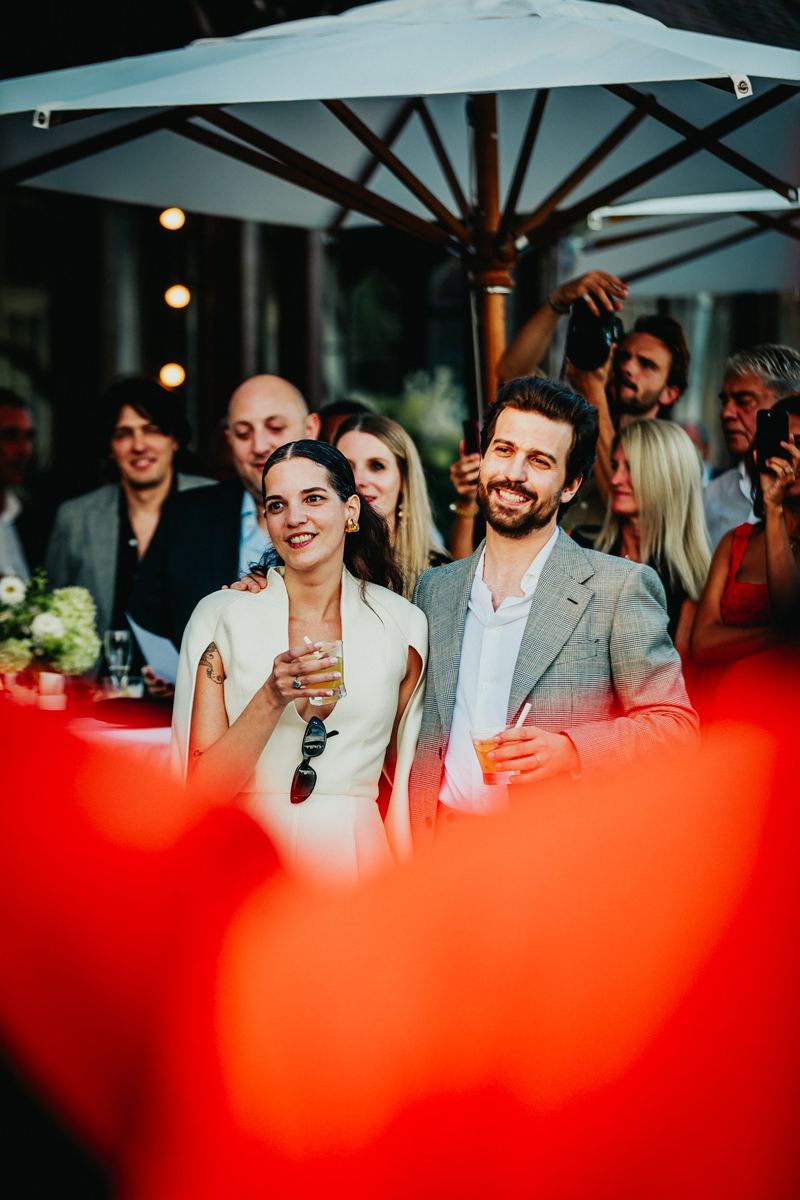 Les mariés au vin d’honneur, aperçus derrière un massif de fleurs rouges
