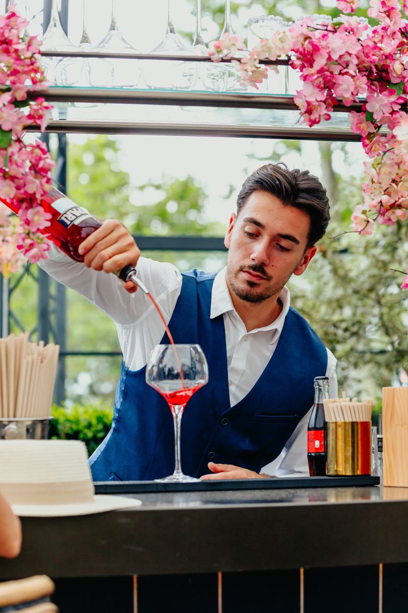 Barman en gilet bleu verse un cocktail sous une treille de fleurs