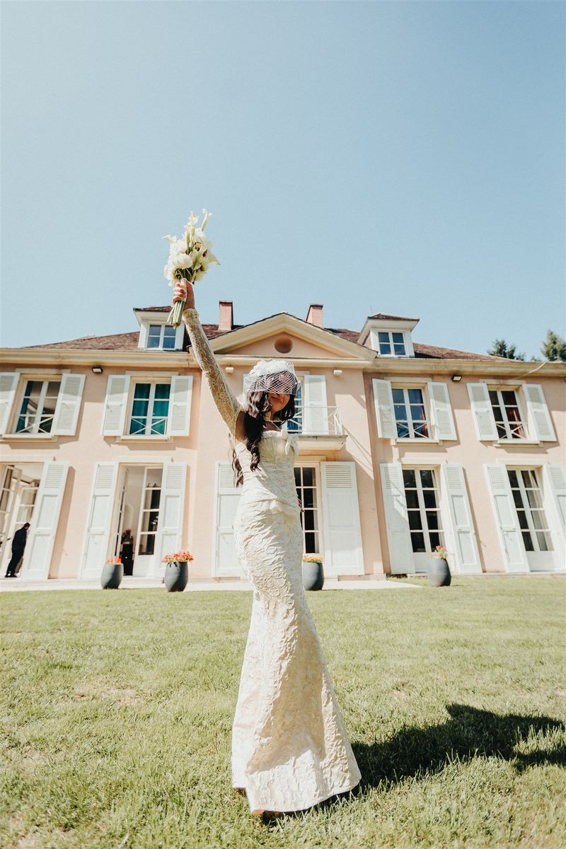 La mariée brandit son bouquet devant la façade rose d’une demeure de réception