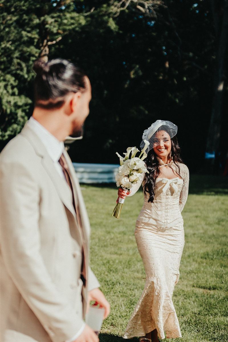 Découverte de la mariée : elle s’avance, bouquet à la main, le marié flou au premier plan